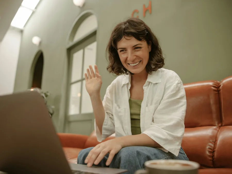 A woman on her sofa waving to her practitioner on a laptop